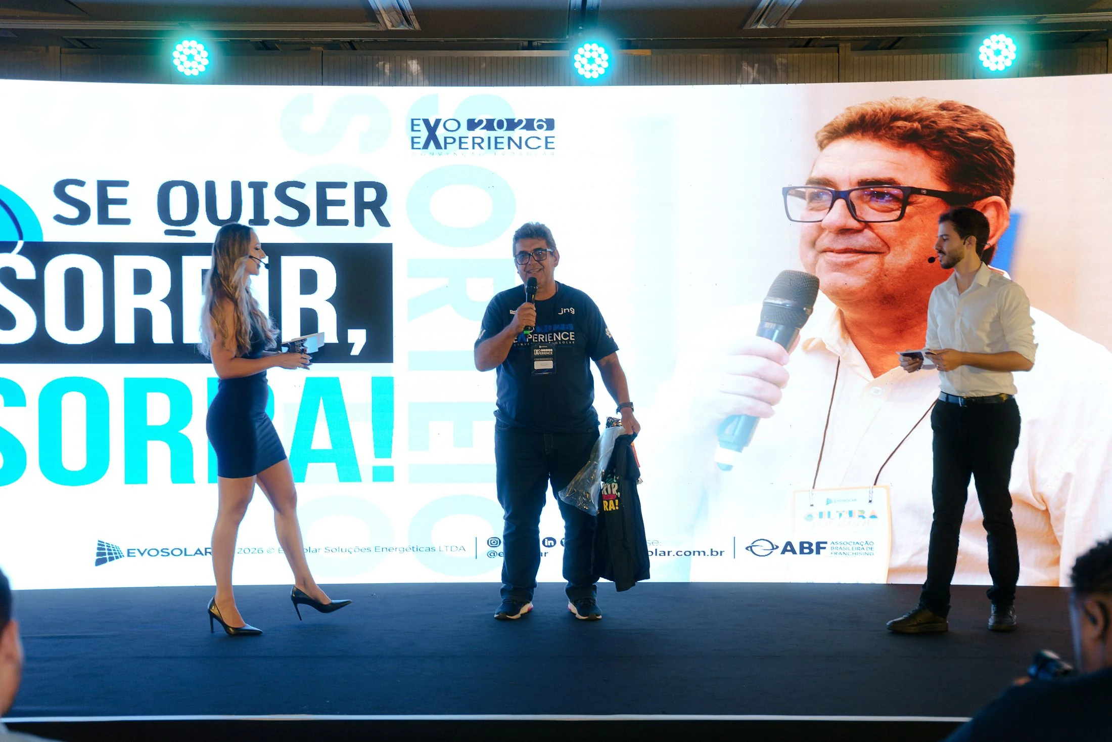Sorteio de brindes no palco da convenção de 2026, diante do telão “Se quiser sorrir, sorra!”